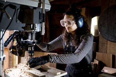 Frau mit langen braunen Haaren, die eine Latzhose, eine Schutzbrille und einen Gehörschutz trägt, steht in einer Holzwerkstatt und benutzt eine elektrische Bohrmaschine. - MINF14460