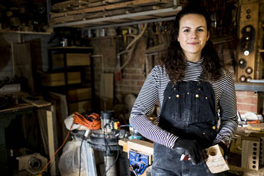 Woman with long brown hair wearing dungarees standing in wood workshop, smiling at camera. - MINF14464