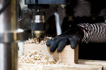 Nahaufnahme einer Person mit Schutzhandschuhen bei der Arbeit mit einer elektrischen Bohrmaschine in einer Holzwerkstatt. - MINF14470
