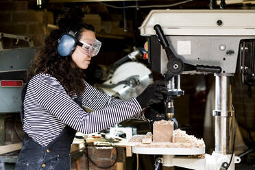 Frau mit langen braunen Haaren, die eine Latzhose, eine Schutzbrille und einen Gehörschutz trägt, steht in einer Holzwerkstatt und benutzt eine elektrische Bohrmaschine. - MINF14472