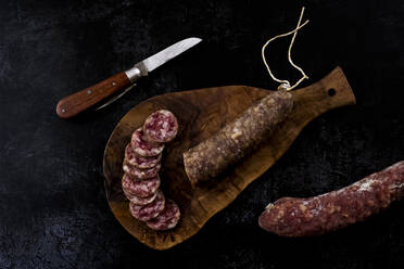 High angle close up of knife, sliced salami on wooden cutting board on black background. - MINF14492