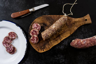 High angle close up of knife, sliced salami on wooden cutting board and white enamel plate on black background. - MINF14494