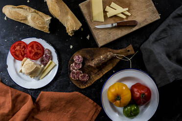 High angle close up of a selection of cheeses, tomatoes, salami and French baguette on white enamel plates on black background. - MINF14495