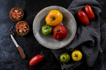 Nahaufnahme eines Messers, eines grauen Tellers und eines Tuchs sowie einer Auswahl frischer Tomaten auf schwarzem Hintergrund. - MINF14496