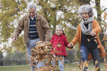 Playful grandparents and granddaughter kicking autumn leaves in park - CAIF24513