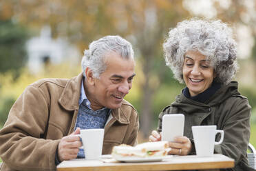 Senior couple with smart phone, eating lunch and drinking coffee in park - CAIF24542