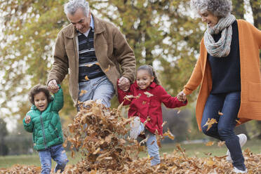 Verspielte Großeltern und Enkelkinder beim Kicken von Herbstblättern im Park - CAIF24547