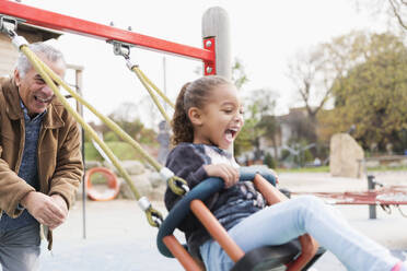 Playful grandfather pushing granddaughter on playground swing - CAIF24587