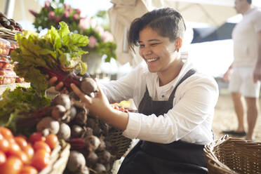 Smiling young woman working, arranging produce at farmer’s market - CAIF24608