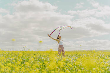 Glückliche junge Frau, die sich mit bunten Bändern auf einer Blumenwiese im Frühling bewegt - ERRF02879