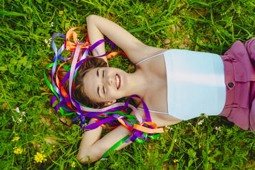 Happy young woman with colourful ribbons lying in a meadow - ERRF02897