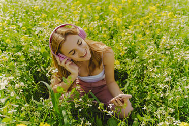 Happy young woman with headphones and smartphone in a flower meadow in spring - ERRF02930