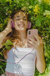 Happy young woman with headphones and smartphone lying in a flower meadow in spring - ERRF02932