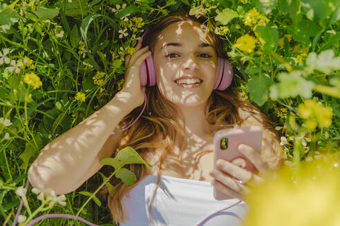 Porträt einer glücklichen jungen Frau mit Kopfhörern und Smartphone auf einer Blumenwiese im Frühling - ERRF02933