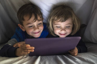 Portrait of happy brother and little sister lying side by side under a blanket using digital tablet - JRFF04237