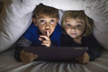 Porträt eines Bruders und einer kleinen Schwester, die Seite an Seite unter einer Decke liegen und ein digitales Tablet benutzen - JRFF04239