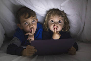Porträt eines Bruders und einer kleinen Schwester, die Seite an Seite unter einer Decke liegen und ein digitales Tablet benutzen - JRFF04240