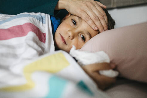 Portrait of sick boy lying in bed while his mother touching his forehead - JRFF04243