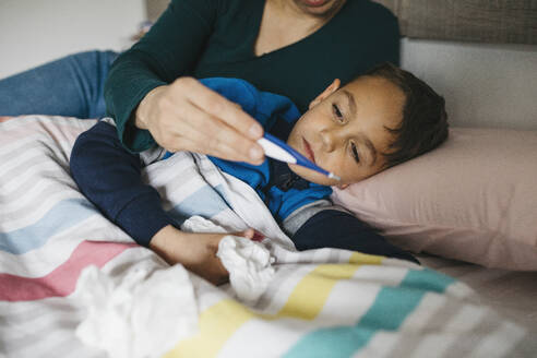 Sick boy with tissue in his hand lying in bed looking together with his mother at digital thermometer - JRFF04244