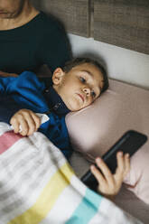Porträt eines kranken kleinen Jungen, der im Bett liegt und auf sein Smartphone schaut - JRFF04245