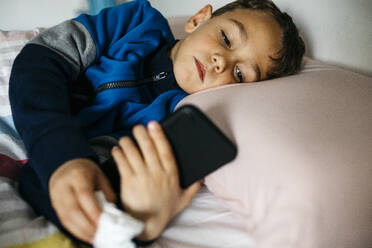 Porträt eines kranken kleinen Jungen, der im Bett liegt und auf sein Smartphone schaut - JRFF04246