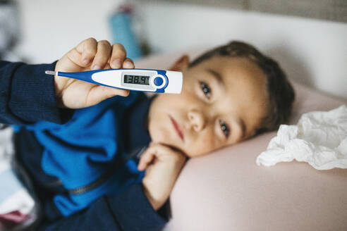 Porträt eines kranken kleinen Jungen, der im Bett liegt und ein digitales Thermometer zeigt - JRFF04247
