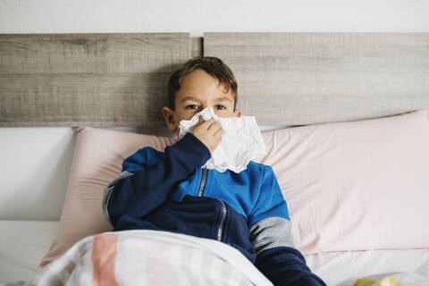 Porträt eines kranken Jungen, der im Bett liegt und sich die Nase putzt, lizenzfreies Stockfoto