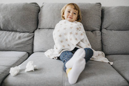 Porträt eines kranken kleinen Mädchens, das zu Hause auf der Couch sitzt und in eine Decke eingewickelt ist - JRFF04250