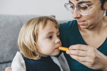 Portrait of sick little girl sitting on couch while mother giving her medication - JRFF04252