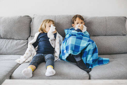 Little girl and her brother sitting side by side on the couch at home blowing noses - JRFF04257