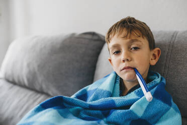 Portrait of sick boy sitting on couch with digital thermometer in his mouth - JRFF04259