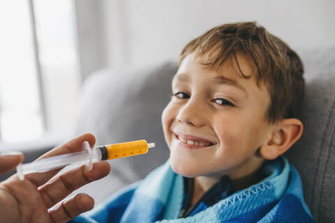 Porträt eines grinsenden kranken Jungen, der auf der Couch sitzt, während seine Mutter ihm Medikamente gibt, lizenzfreies Stockfoto