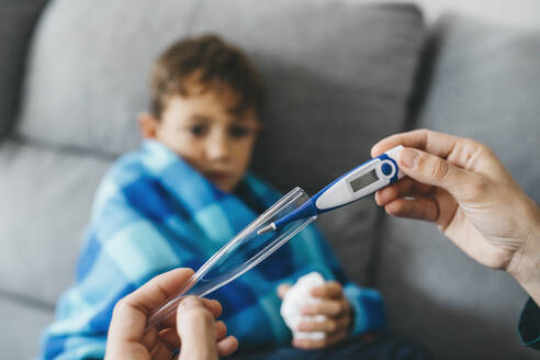 Die Hand der Mutter nimmt ein digitales Thermometer, während ihr Sohn auf der Couch im Hintergrund wartet - JRFF04261