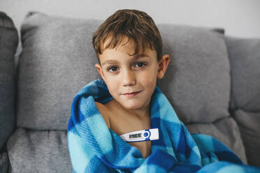 Portrait of sick boy sitting on couch with digital thermometer - JRFF04263