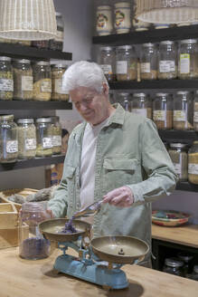 Älterer Mann hinter dem Tresen seines Ladens, der getrocknete Lavendelblüten in eine Waage schüttet - AFVF05861