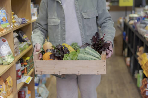 Nahaufnahme eines älteren Mannes, der eine Kiste mit Gemüse in einem kleinen Lebensmittelgeschäft trägt - AFVF05880