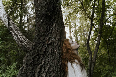 Young redhaired woman hugging tree trunk in forest - AFVF05928
