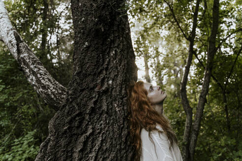 Junge rothaarige Frau umarmt Baumstamm im Wald - AFVF05928