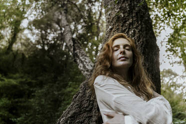 Junge rothaarige Frau mit geschlossenen Augen lehnt sich an einen Baumstamm - AFVF05929