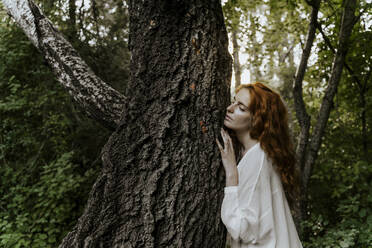 Young redhaired woman hugging tree trunk in the forest - AFVF05931