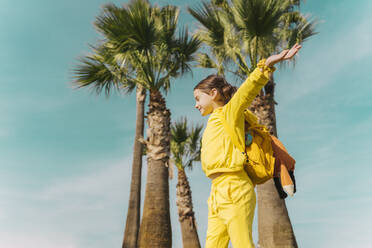 Smiling little girl with backpack standing in front of palms - ERRF02936