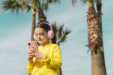 Portrait of little girl wearing yellow jacket listening music with headphones looking at smartphone - ERRF02942