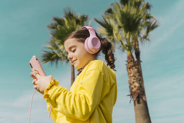 Portrait of smiling little girl wearing yellow jacket listening music with headphones looking at smartphone - ERRF02943