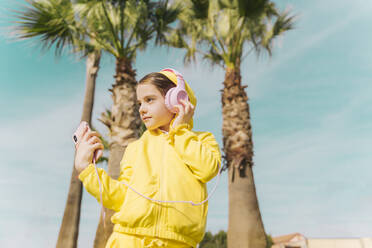 Portrait of little girl wearing yellow hooded jacket listening music with headphones and smartphone - ERRF02946