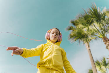 Portrait of little girl wearing yellow hooded jacket listening music with headphones - ERRF02948