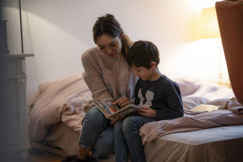 Mutter und kleiner Sohn sitzen zusammen auf dem Bett und schauen sich ein Bilderbuch an - KNSF07937