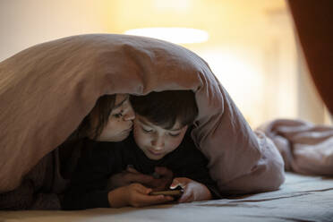 Mutter und kleiner Sohn liegen zusammen auf einem Bett mit Decke und schauen einen Film auf dem Smartphone - KNSF07941