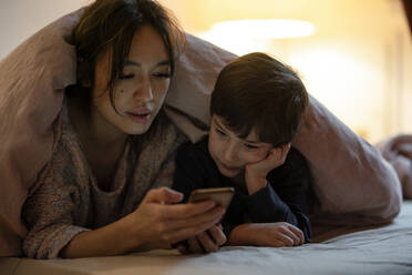 Mutter und kleiner Sohn liegen zusammen auf einem Bett mit Decke und schauen einen Film auf dem Smartphone - KNSF07942