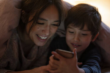 Mother and little son lying together on bed covered with blanket watching movie on smartphone - KNSF07943