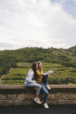 Zwei junge Frauen sitzen auf einer Mauer in einer ländlichen Landschaft und machen ein Selfie, Greve in Chianti, Toskana, Italien, lizenzfreies Stockfoto